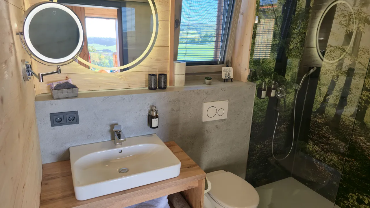 Modern chalet bathroom with wooden vanity, white washbasin, illuminated round mirror and walk-in shower with forest motif