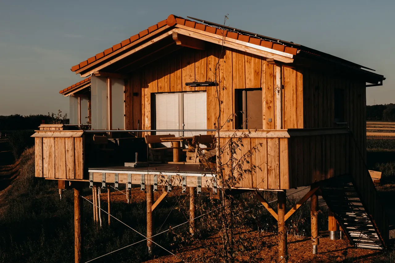 Wooden chalet on stilts with red tiled roof, outdoor staircase and terrace with rustic seating in golden evening light
