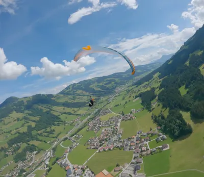 Aerial view of a solo paraglider flying high above a green alpine valley, with a village, road, and forested mountainside spread out below