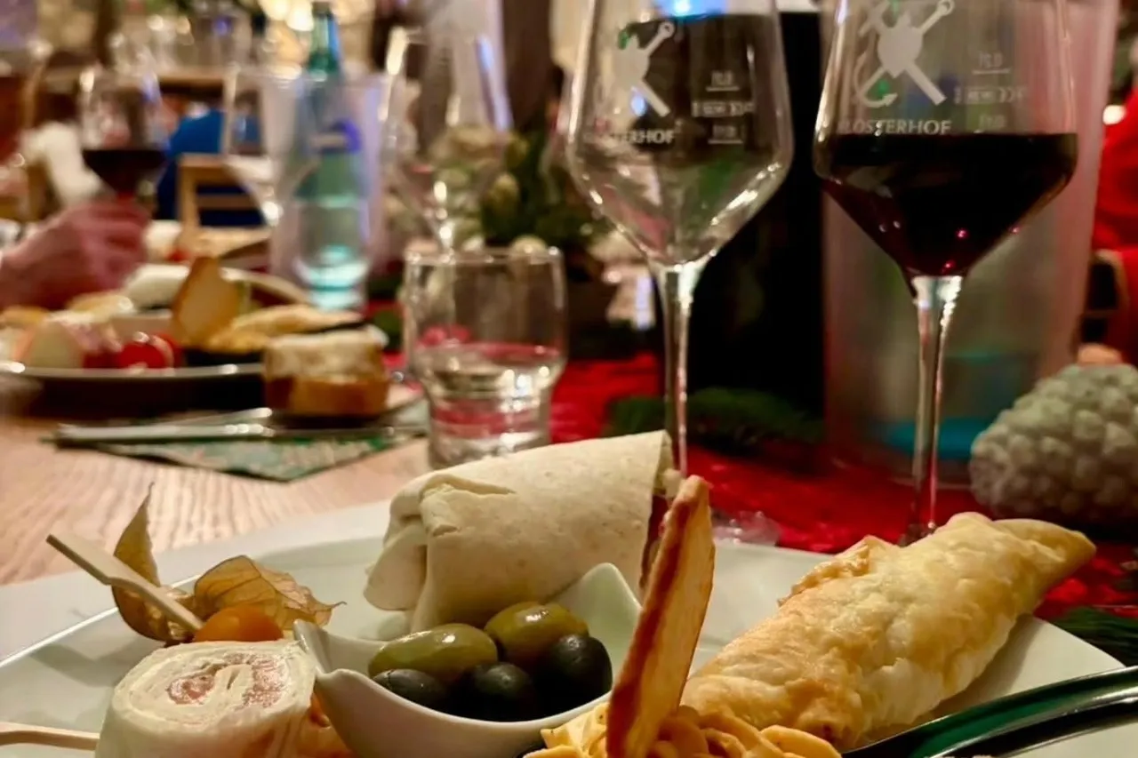 Plate of finger food with olives, wraps, cheese and tomatoes next to wine glasses while the host presents in the background