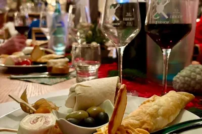 Plate of finger food with olives, wraps, cheese and tomatoes next to wine glasses while the host presents in the background