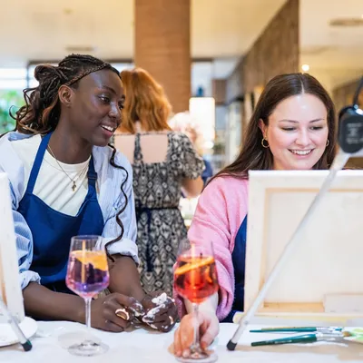 Two women sitting next to each other drinking Aperol Spritz and painting