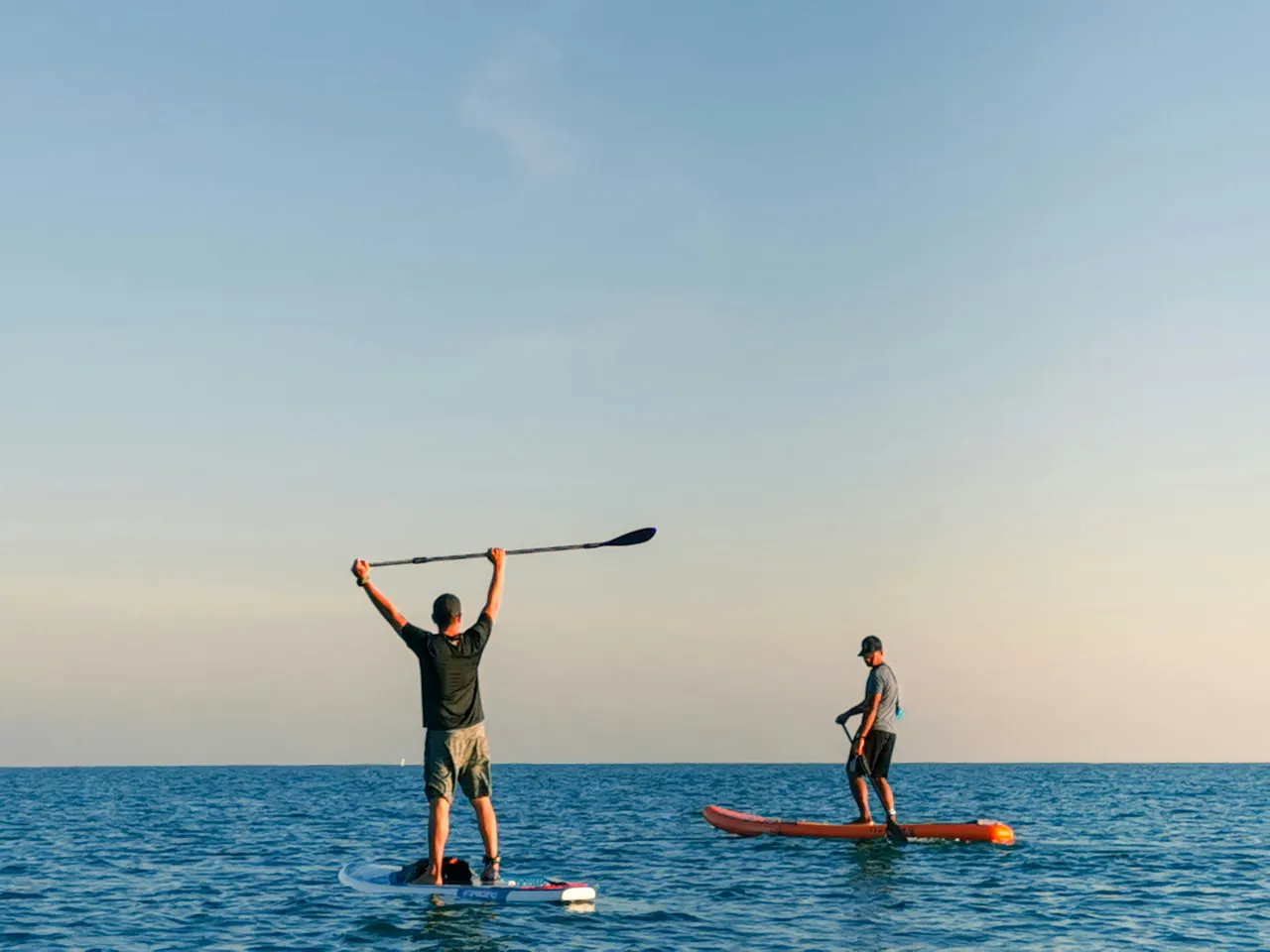 Two people on stand-up paddleboards in the open ocean under a clear sky, with one person holding their paddle high above their head in celebration.