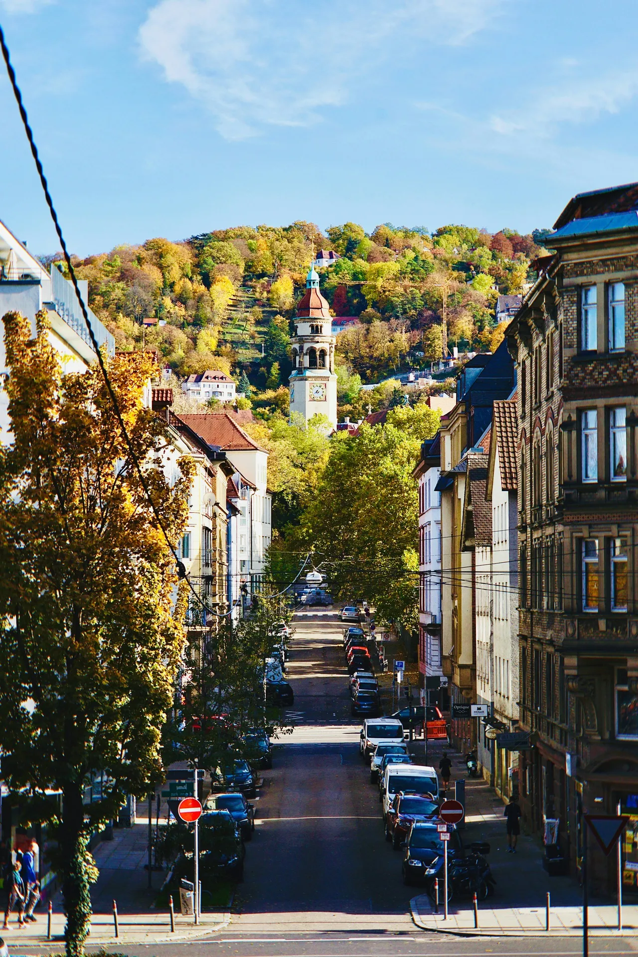 City street in Stuttgart lined with parked cars, leading toward a hillside church tower in autumn