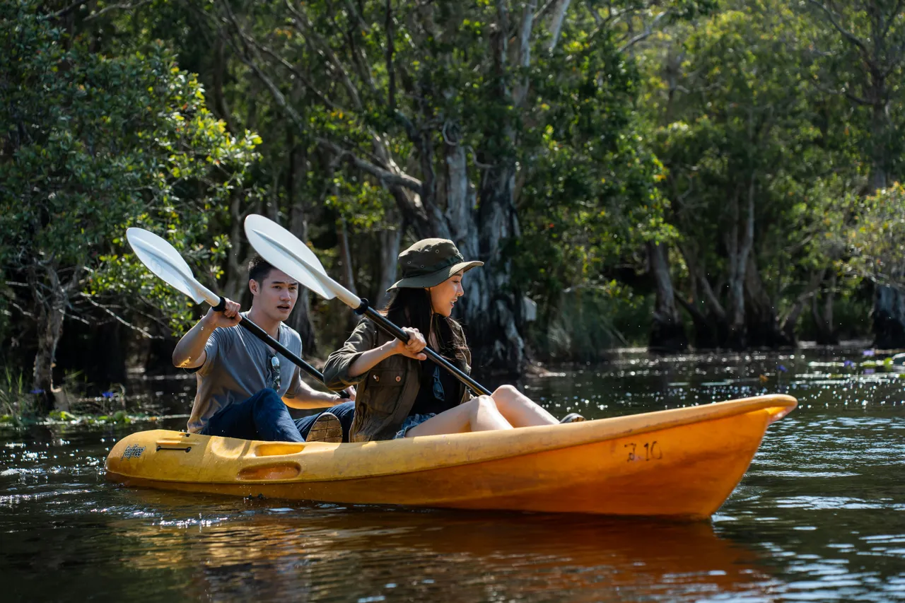 A young man and a woman in a sun hat navigate a yellow tandem kayak through a scenic wetland area surrounded by trees.