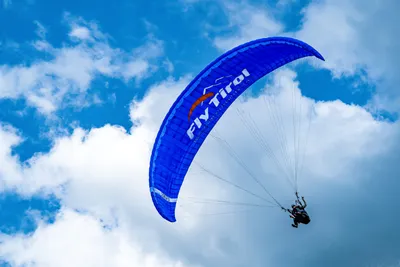 A blue Fly Tirol tandem paraglider canopy soaring against a bright blue sky with white clouds, with the pilot and passenger visible below