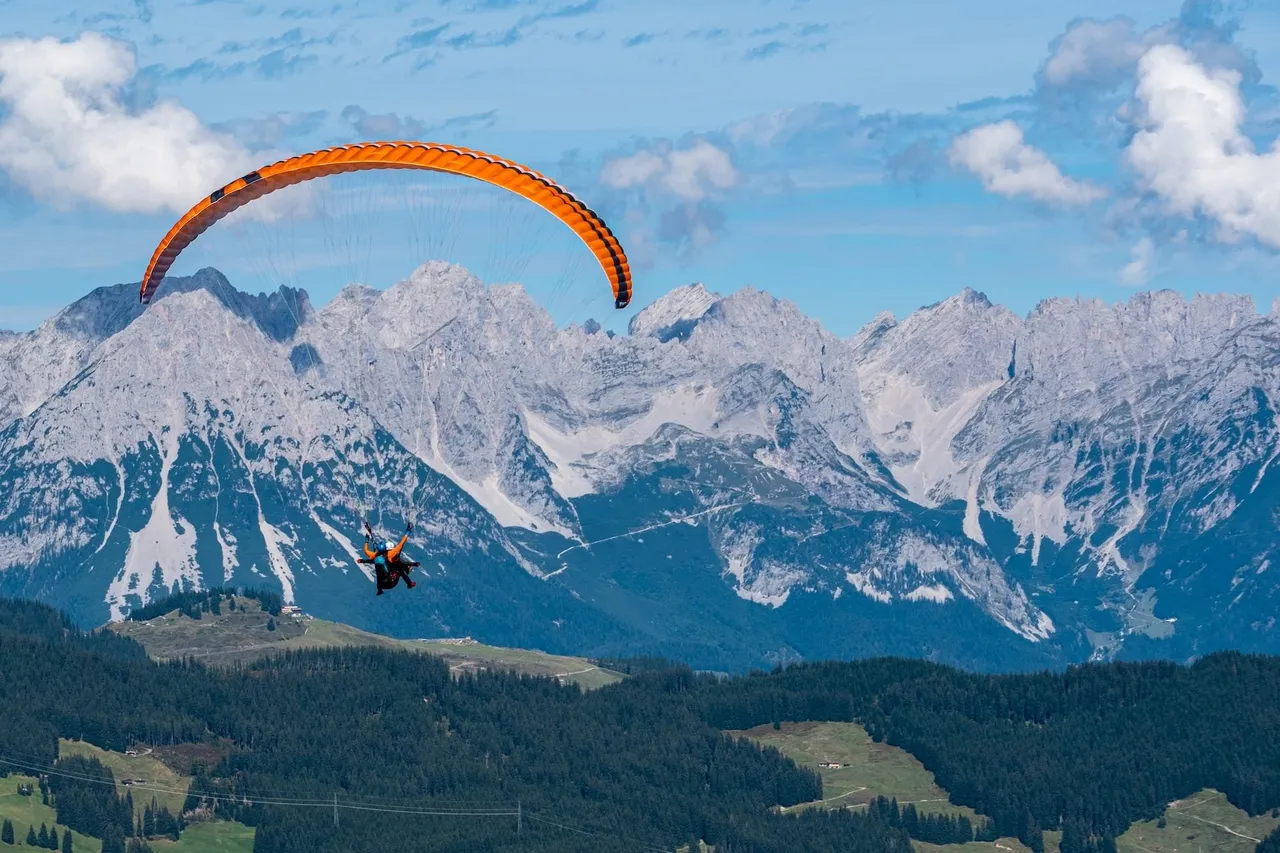 An orange tandem paraglider gliding in front of the rugged, snow-streaked peaks of the Wilder Kaiser mountain range in Tyrol, with green forests below