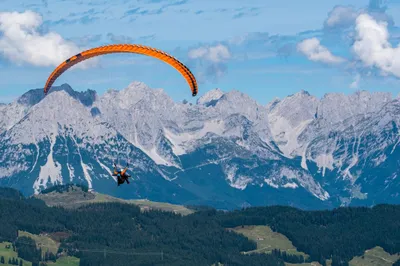 An orange tandem paraglider gliding in front of the rugged, snow-streaked peaks of the Wilder Kaiser mountain range in Tyrol, with green forests below
