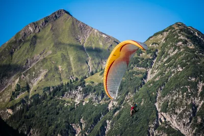Tandem paraglider with orange wing flying past green alpine peaks in Tyrol