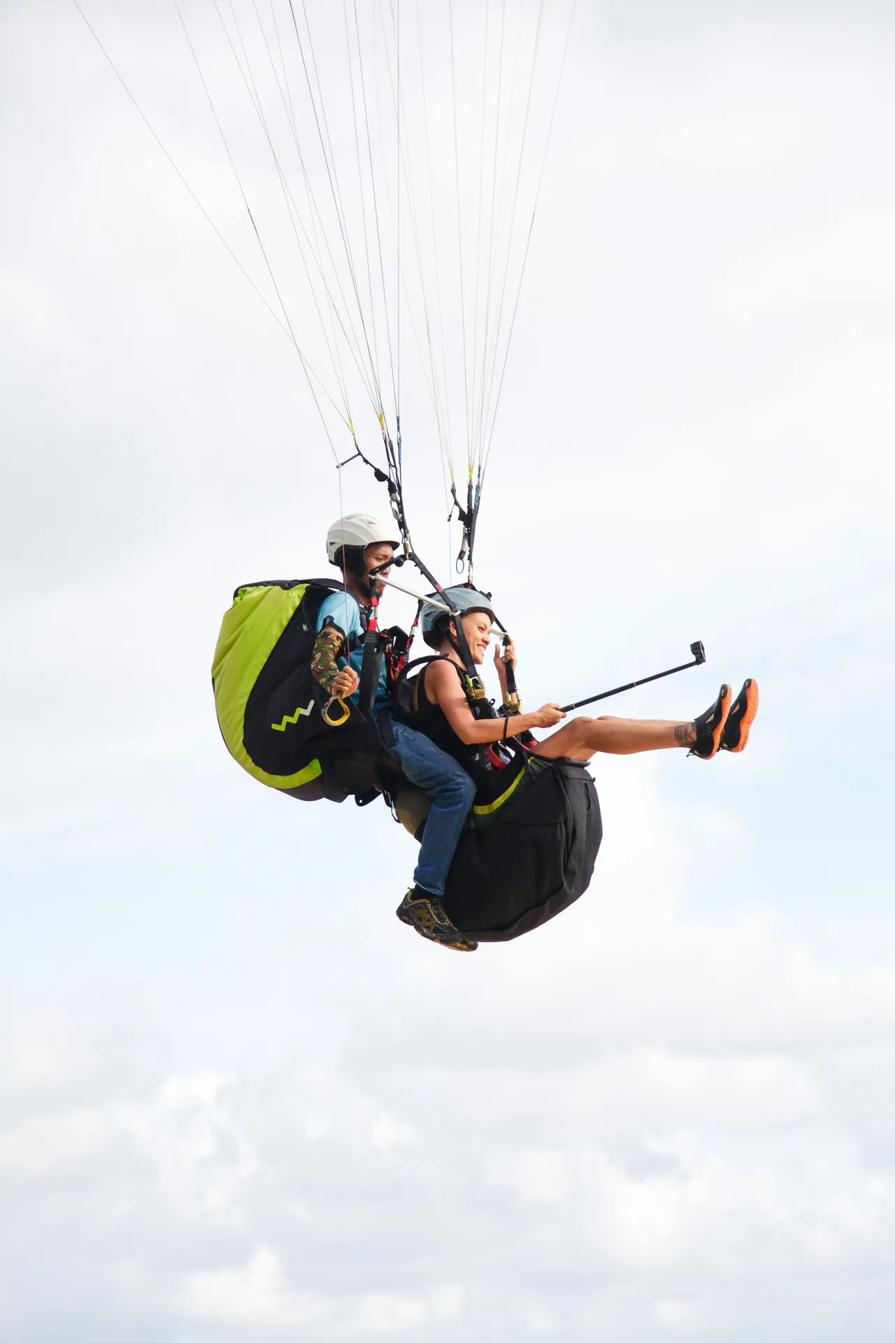 A close-up side view of two people in helmets paragliding tandem against a bright, cloudy white sky, with the passenger holding out a selfie stick.