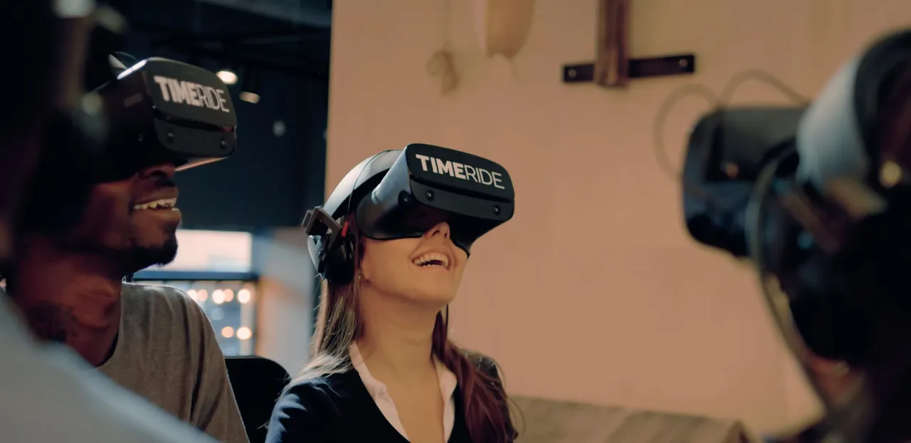 Smiling woman wearing a TimeRide VR headset, laughing with another guest also wearing a headset during a group virtual reality experience