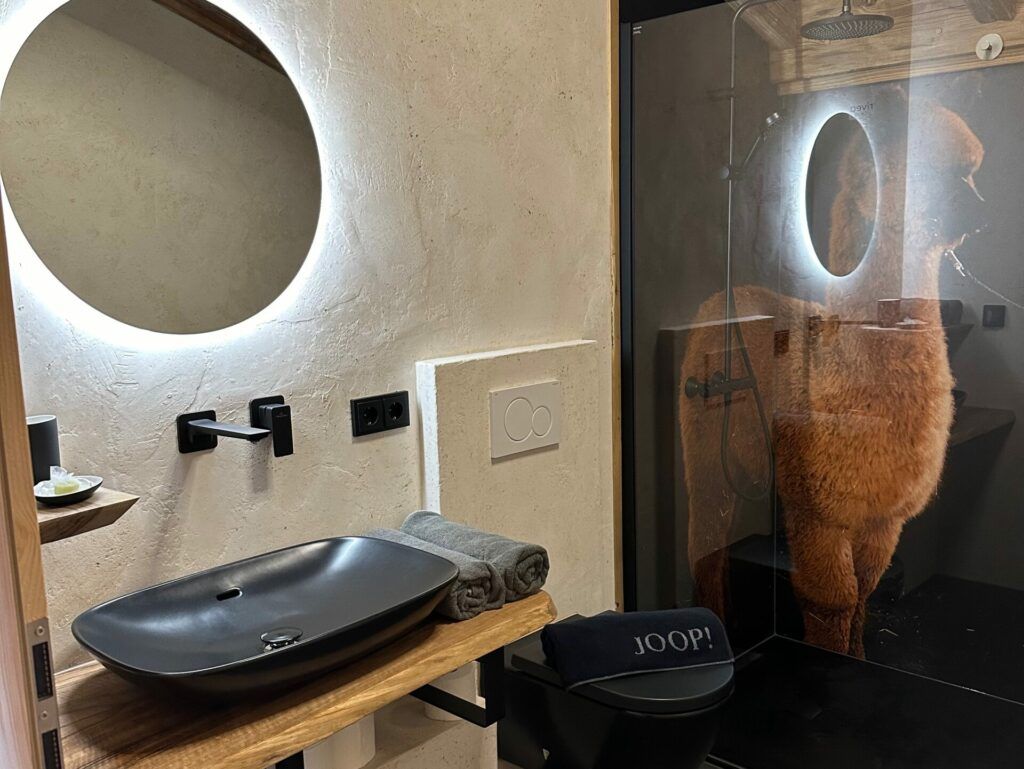Modern treehouse bathroom with round illuminated mirror, black vessel sink on a wooden shelf and glass walk-in shower