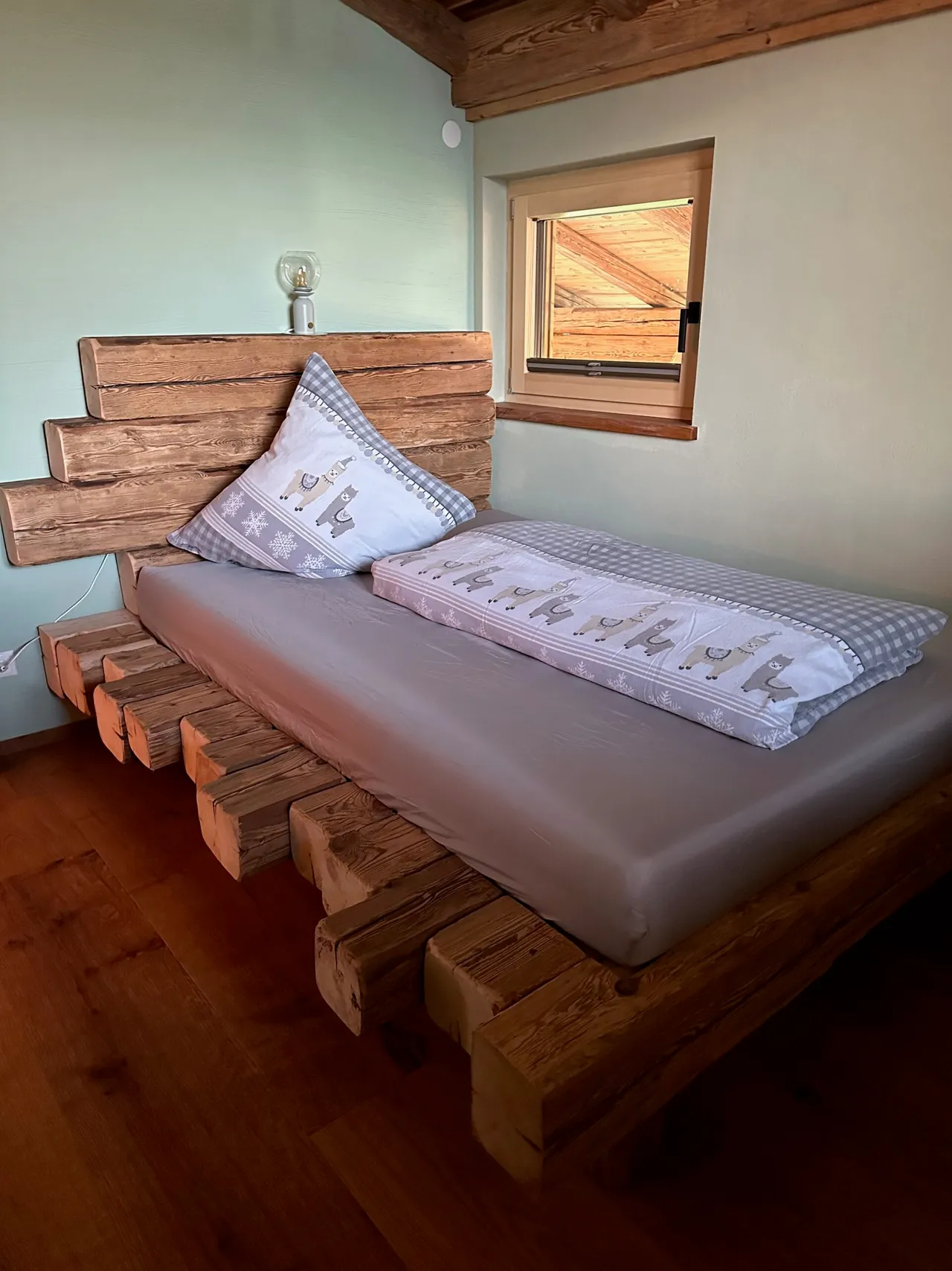 Bed built from reclaimed timber beams with alpaca-print bedding next to a small window in the treehouse