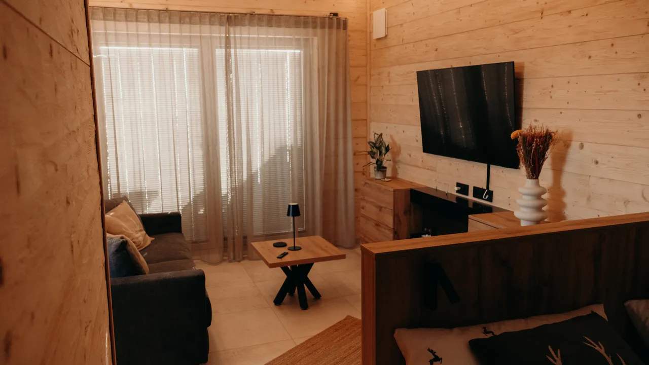 Chalet living room with sofa, small coffee table, wall-mounted TV and sheer curtains in soft evening light