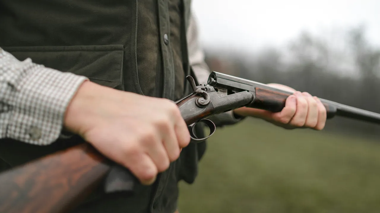 A close-up view of a person's hands holding an antique break-action shotgun with a wooden stock, wearing a green hunting vest and plaid shirt outdoors.