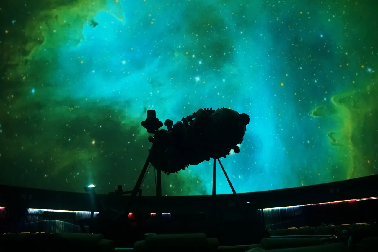 Silhouette of a large mechanical star projector in a planetarium against a bright green and teal nebula projection on the domed screen.
