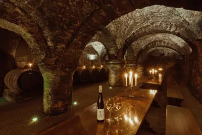 Historic stone wine cellar interior with barrel-vaulted arches, illuminated wooden wine barrels, lit candelabras, and a bottle of wine with filled glasses on a dark table in the foreground.