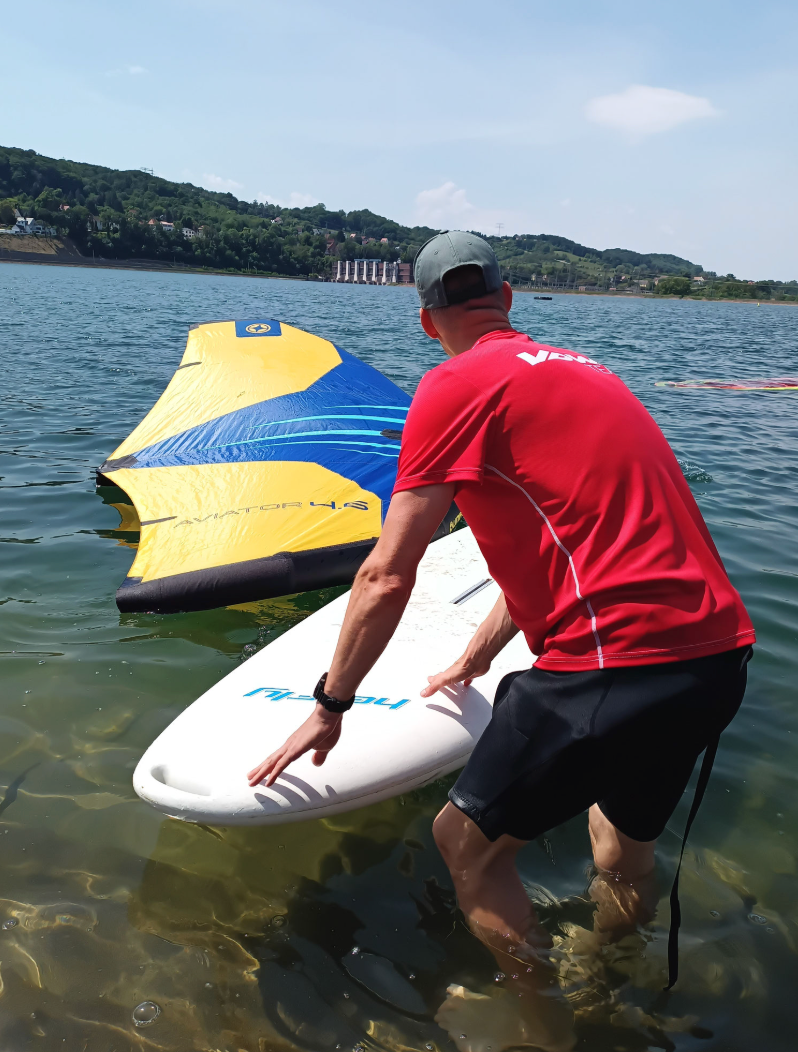 A person putting a windsurf board in the water with a blue and yellow hydrofoil