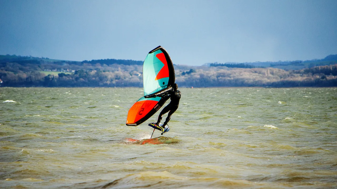 A person wing foiling on a choppy body of water with a rolling green landscape in the background