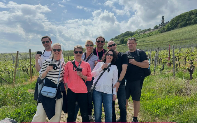 Group of hikers with wine glasses posing in the vineyards above Rüdesheim, with the Niederwald Monument on the hill behind them.