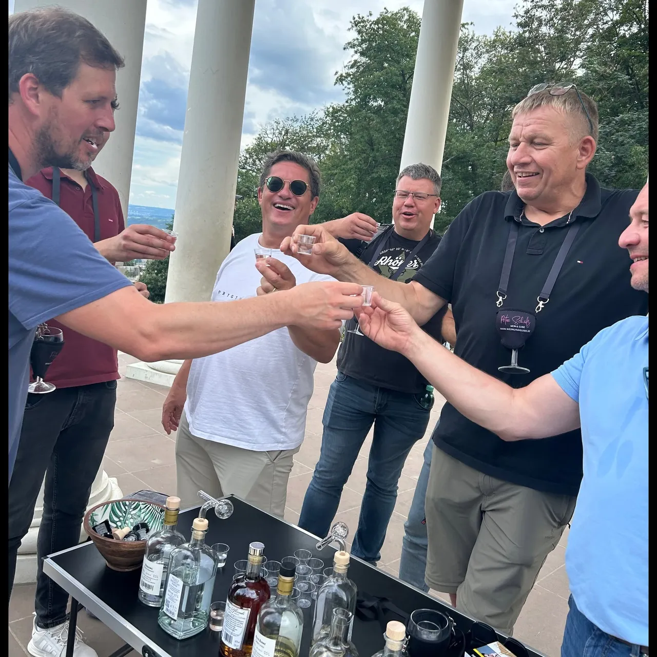Group of men laughing and toasting with small glasses at an outdoor fruit brandy tasting beside white columns.