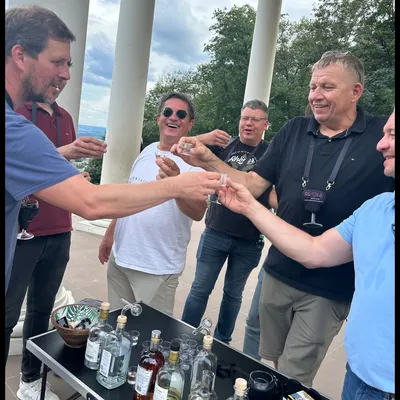 Group of men laughing and toasting with small glasses at an outdoor fruit brandy tasting beside white columns.