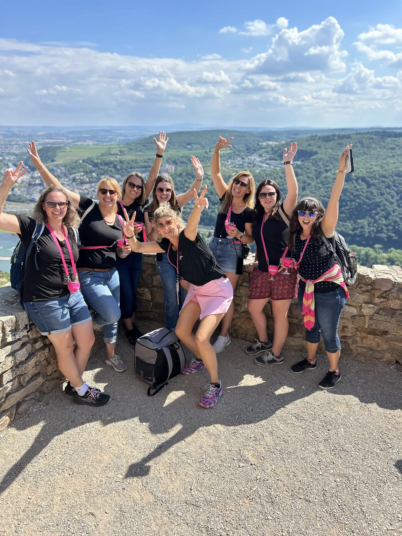 Cheerful group of women with pink wine glasses raising their arms at a viewpoint above the Rhine valley.