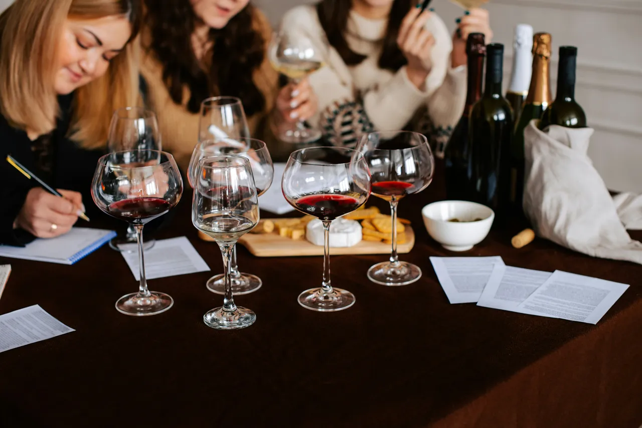 A table set with multiple glasses of red and white wine, wine bottles, and evaluation sheets as participants take notes during a structured wine tasting event.