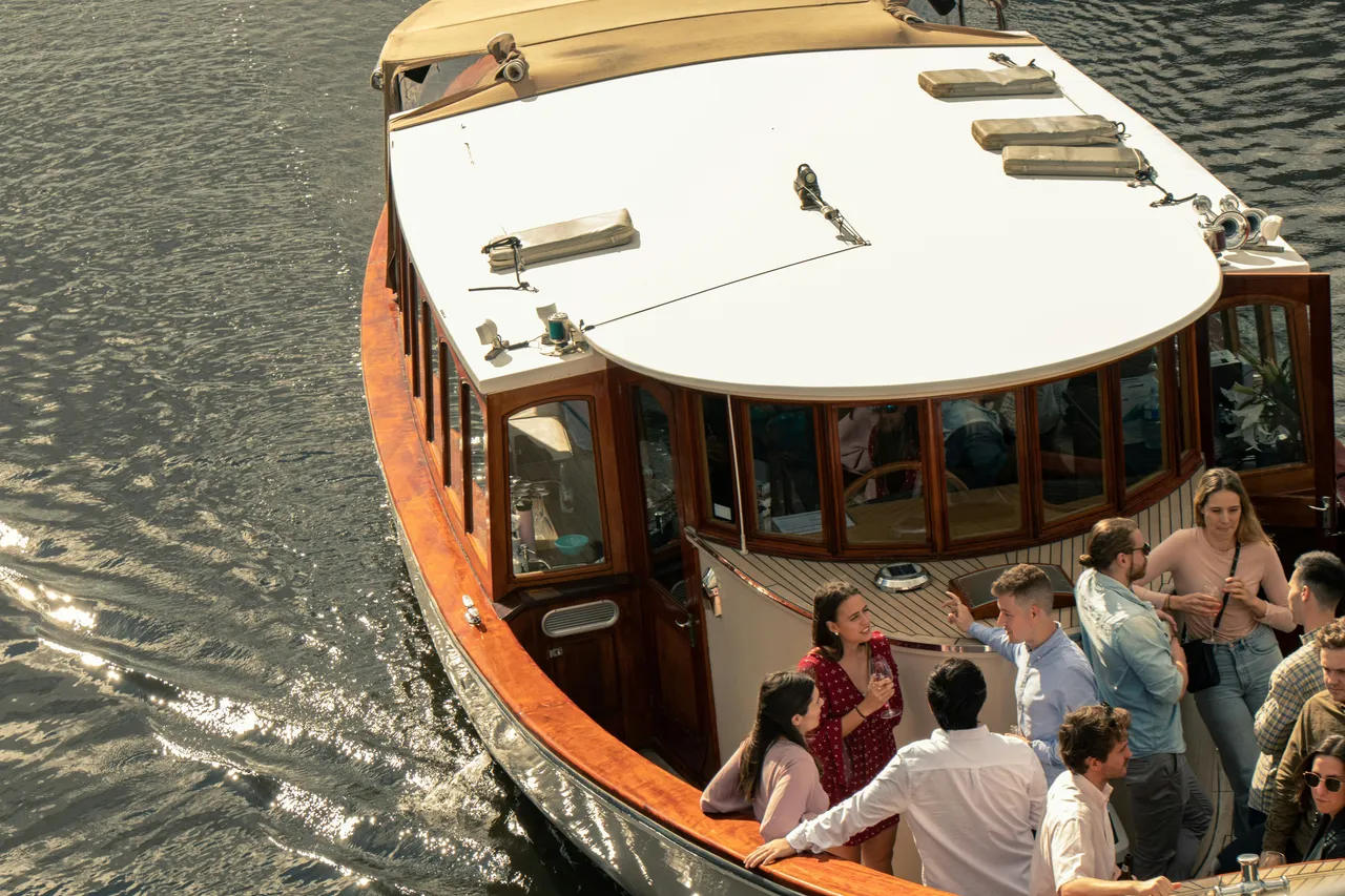 High-angle view of a group of people socializing and chatting on the deck of a classic wooden tour boat on the water.