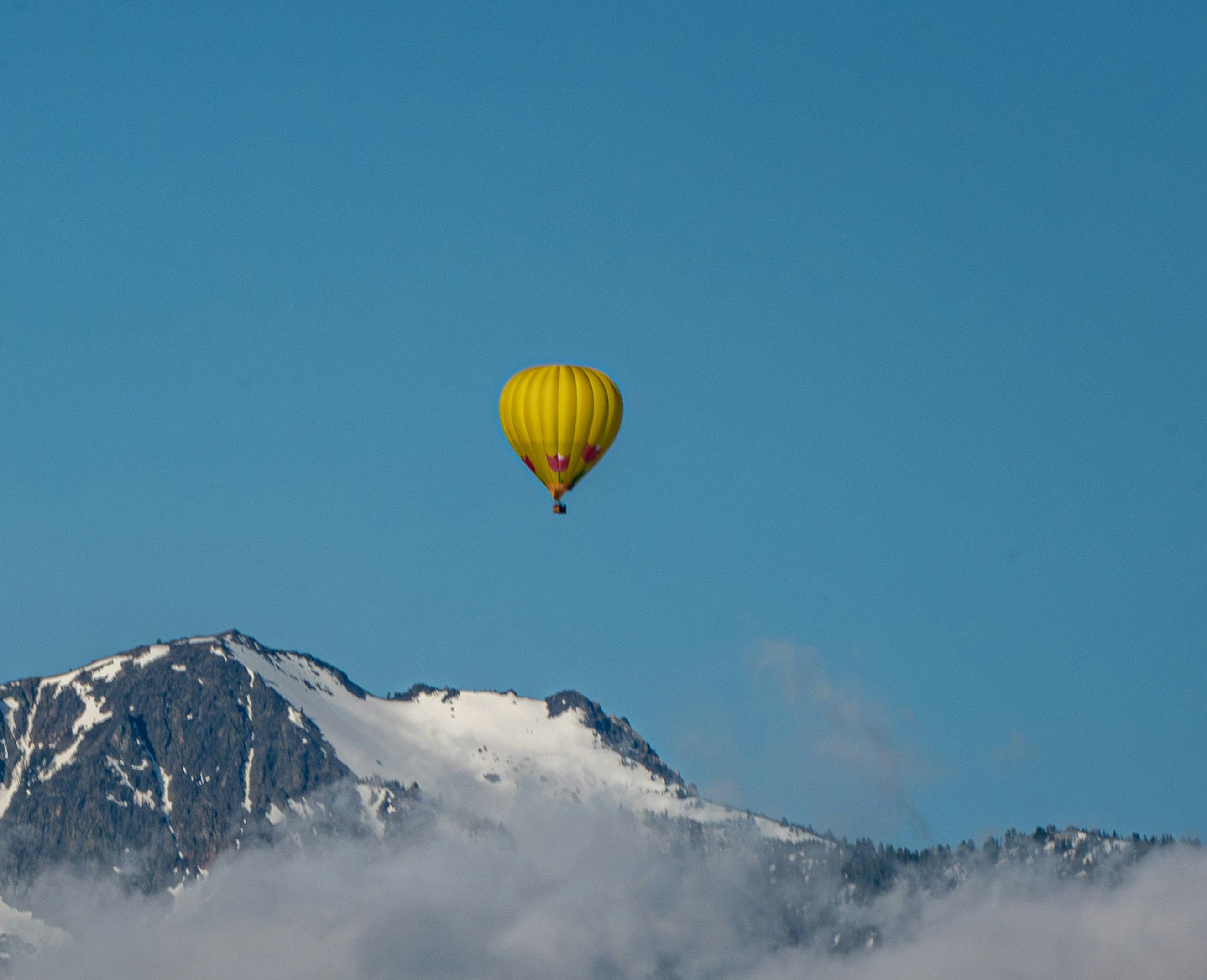 A bright yellow hot air balloon flying high in a blue sky above a snow-patched mountain peak wrapped in clouds.