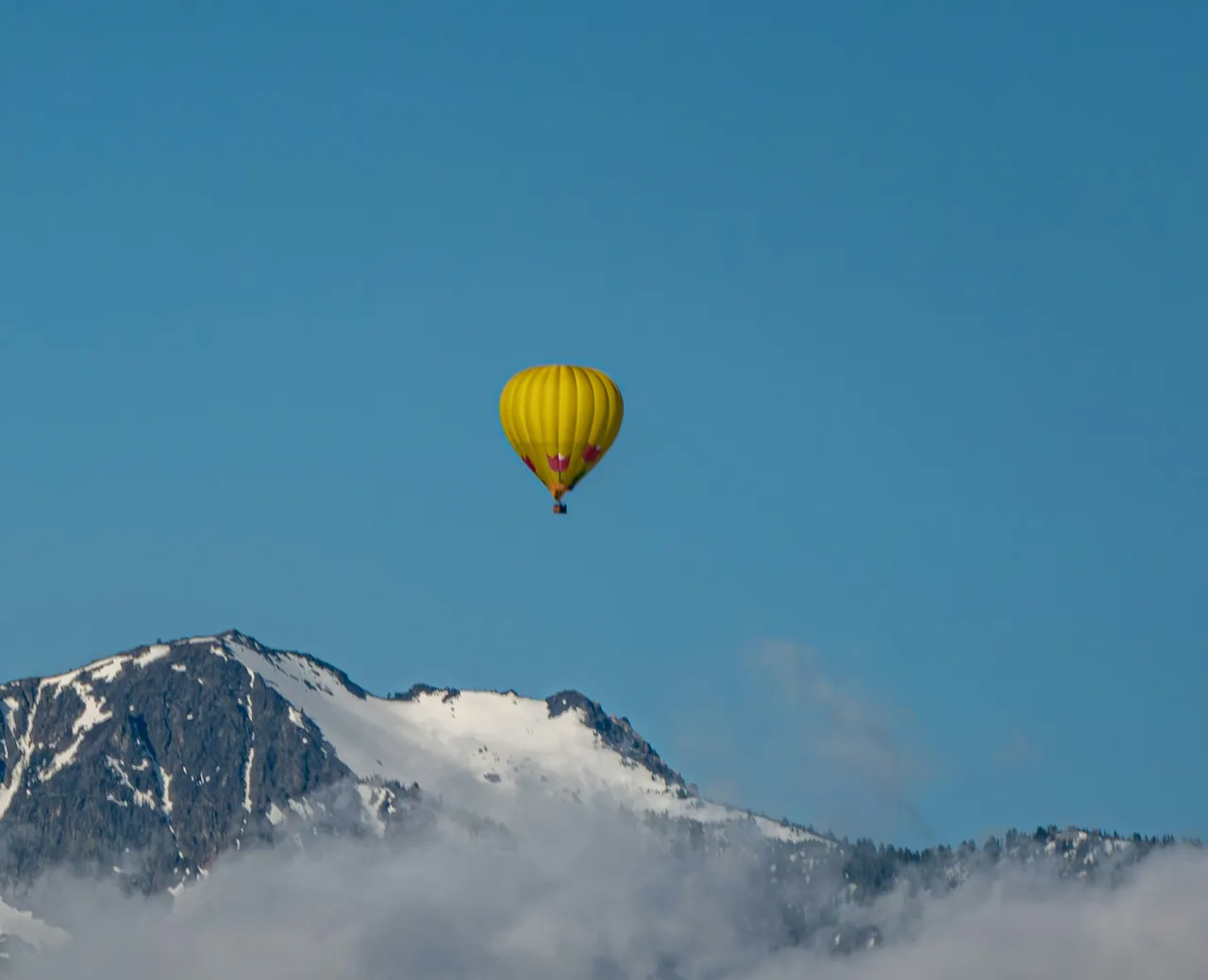 A bright yellow hot air balloon flying high in a blue sky above a snow-patched mountain peak wrapped in clouds.