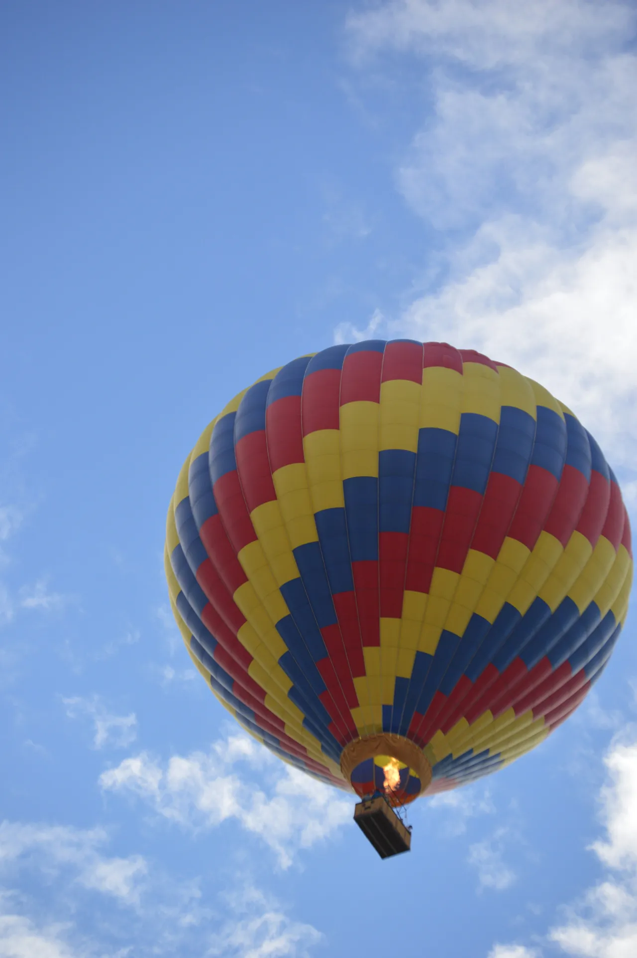 A large hot air balloon with a spiraling checkered pattern of red, yellow, and blue panels floats in a bright blue sky filled with wispy white clouds.